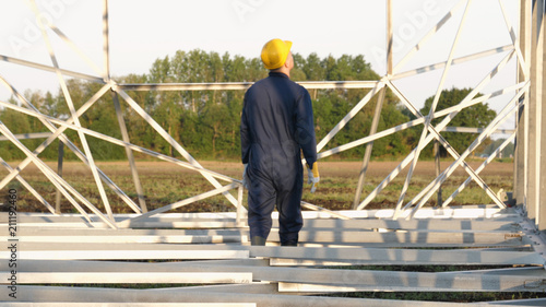 An electrician or builder, an employee in a blue robe, wearing a yellow helmet, is preparing to install electric poles, an electric tower, and energy. © dkHDvideo