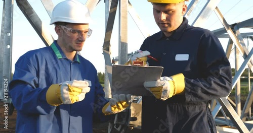 An electrician or builder and engineer, in a blue robe, in a yellow and white helmet, check the technical overview for strength, prepare electric poles, an electric tower, energy, and workers.