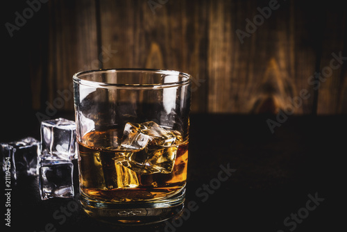Two whiskey shot glasses on dark wooden background, with ice cubes, copy space