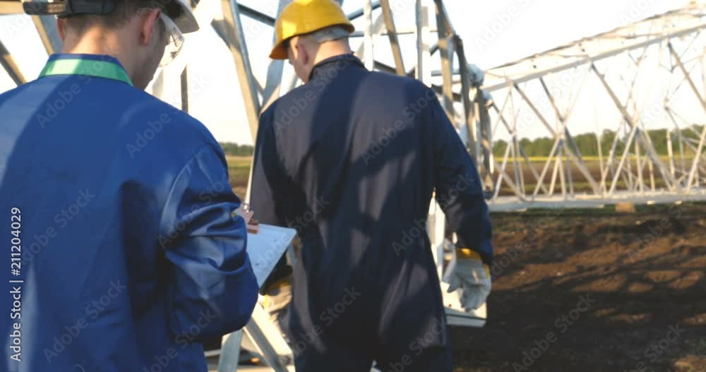 An electrician or builder and engineer, in a blue robe, in a yellow and white helmet, check the technical overview for strength, prepare electric poles, an electric tower, energy, and workers. An electrician or builder and engineer, in a blue robe, in a yellow and white helmet, check the technical overview for strength, prepare electric poles, an electric tower, energy, and workers.