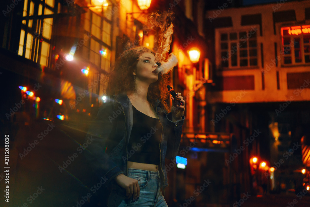 Young attractive red-haired girl smokes electronic cigarette. She walks through small streets of city. Evening city lights blink.