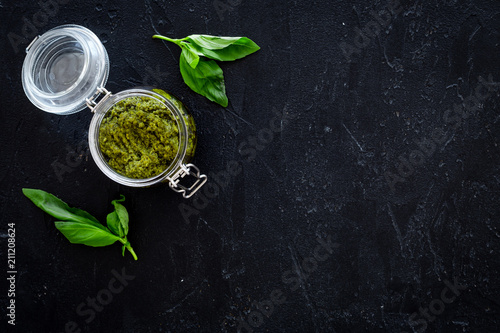 Green pesto sauce in glass jar near basil leaves on black background top view copy space