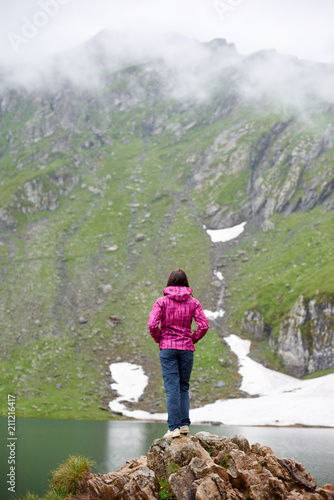 Rear view of young woman hiker with hands in pockets standing on the edge of rocky cliff looking at the blue lake in the mountains in Romania.