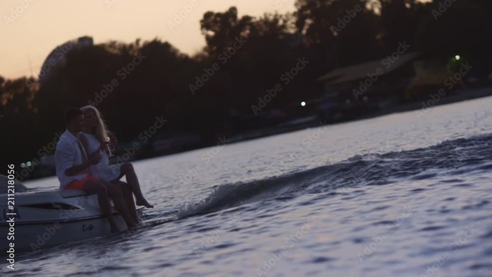 Young people sitting on floating motor boat at sunset. Love couple drinking champagne at yacht. Romantic weekend vacation on river in evening. Lovers celebrating with champagne on boat Young people sitting on floating motor boat at sunset. Love couple drinking champagne at yacht. Romantic weekend vacation on river in evening. Lovers celebrating with champagne on boat