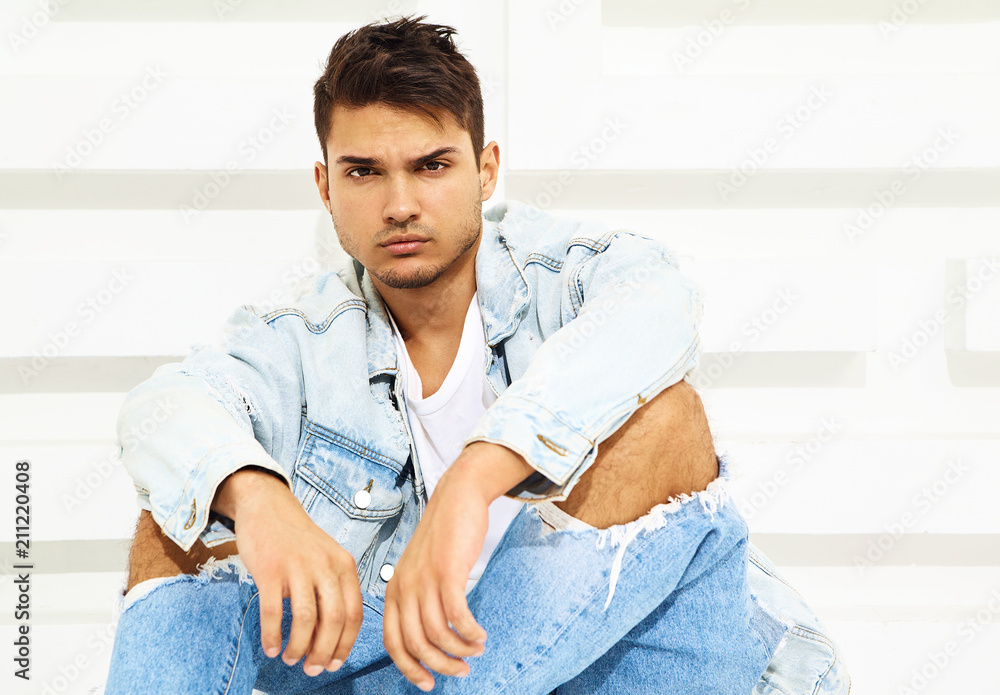 Portrait of handsome young model man dressed in jeans clothes sitting near white textured wall Portrait of handsome young model man dressed in jeans clothes sitting near white textured wall