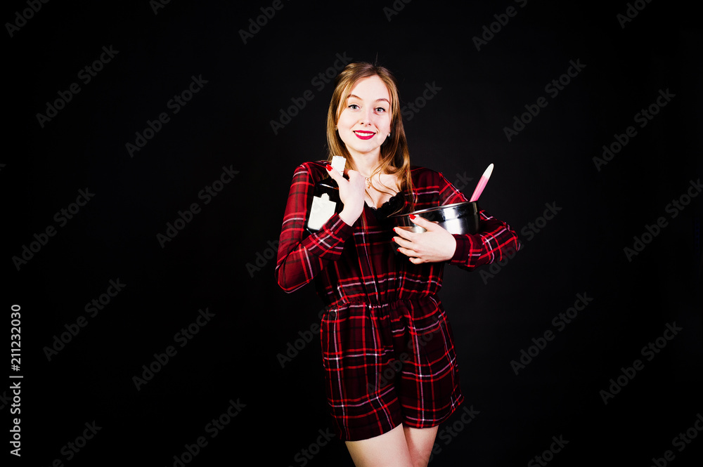 Young housewife in checkered dress with saucepan and kitchen spoon and bottle isolated on black background. Young housewife in checkered dress with saucepan and kitchen spoon and bottle isolated on black background.