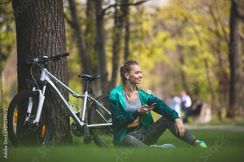 Smiling sporty lady is resting on lawn in park during riding bike. She is sitting and listening to audio tracks on smartphone with earphones