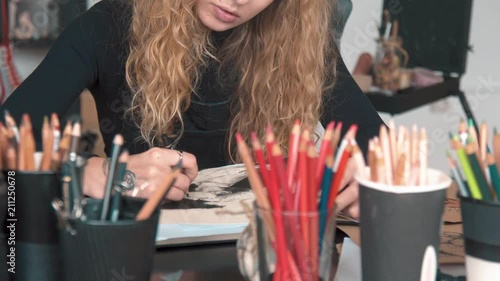 Professional young artist drawing a picture in the sketchbook, working at the desk with lots of various pencils, has lovely tattoo of flower on right hand