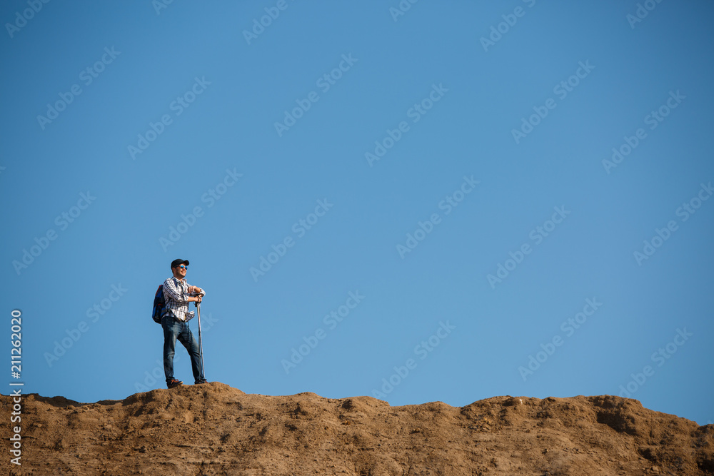 Picture of afar of young tourist man with sticks for walking on hill against blue sky