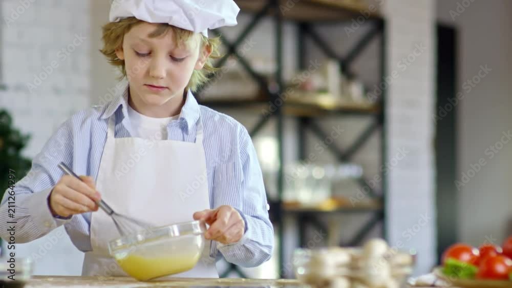 Medium shot of cute little boy in apron and chef hat looking at camera and beating eggs with whisk Medium shot of cute little boy in apron and chef hat looking at camera and beating eggs with whisk