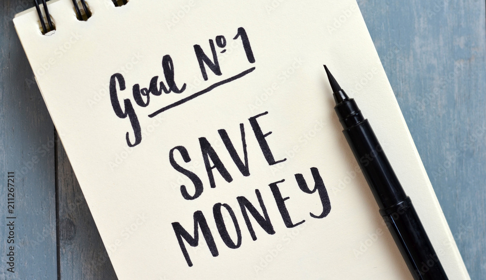 GOAL #1 SAVE MONEY hand-lettered in notebook on blue wooden desk with pen GOAL #1 SAVE MONEY hand-lettered in notebook on blue wooden desk with pen