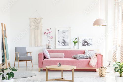 Real photo of a pink couch with pillows standing behind a table, next to an armchair, in front of a shelf with posters and flowers and under a lamp in bright and cozy living room interior