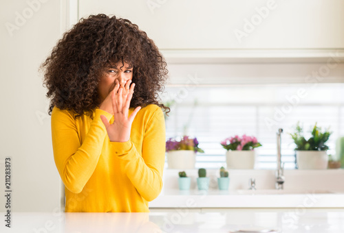 African american woman wearing yellow sweater at kitchen smelling something stinky and disgusting, intolerable smell, holding breath with fingers on nose. Bad smells concept.