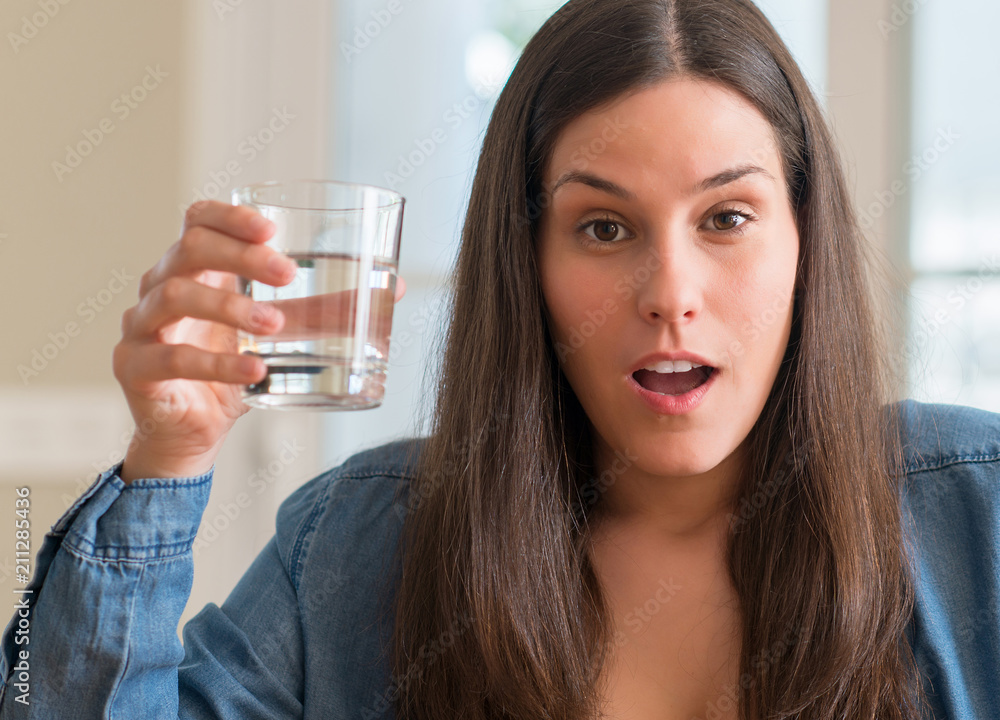 Young woman drinking glass of water at home scared in shock with a surprise face, afraid and excited with fear expression Young woman drinking glass of water at home scared in shock with a surprise face, afraid and excited with fear expression