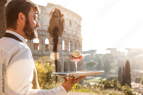 Photography Elegant waiter holding a serving try with a glass of colorful icecream in front