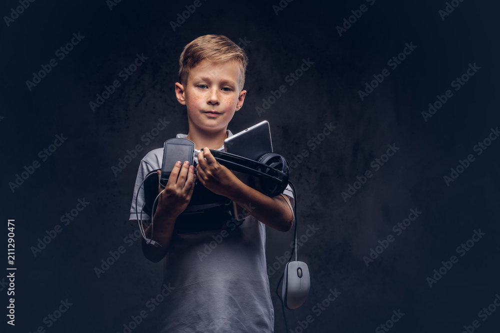 Cute schoolboy dressed in a white t-shirt, holds full digital set for entertainment at a studio. Isolated on dark textured background.