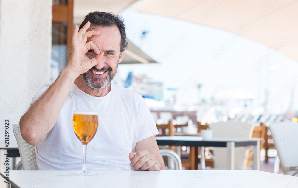 Handsome senior man drinking beer at restaurant with happy face smiling doing ok sign with hand on eye looking through fingers