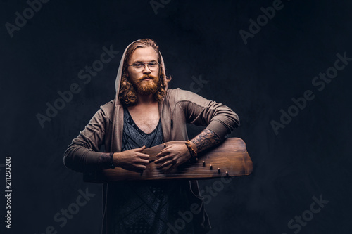 Redhead hipster male with long luxuriant hair and full beard dressed in a hoodie and t-shirt playing on a Russian traditional musical instrument - gusli. Isolated on a dark background.