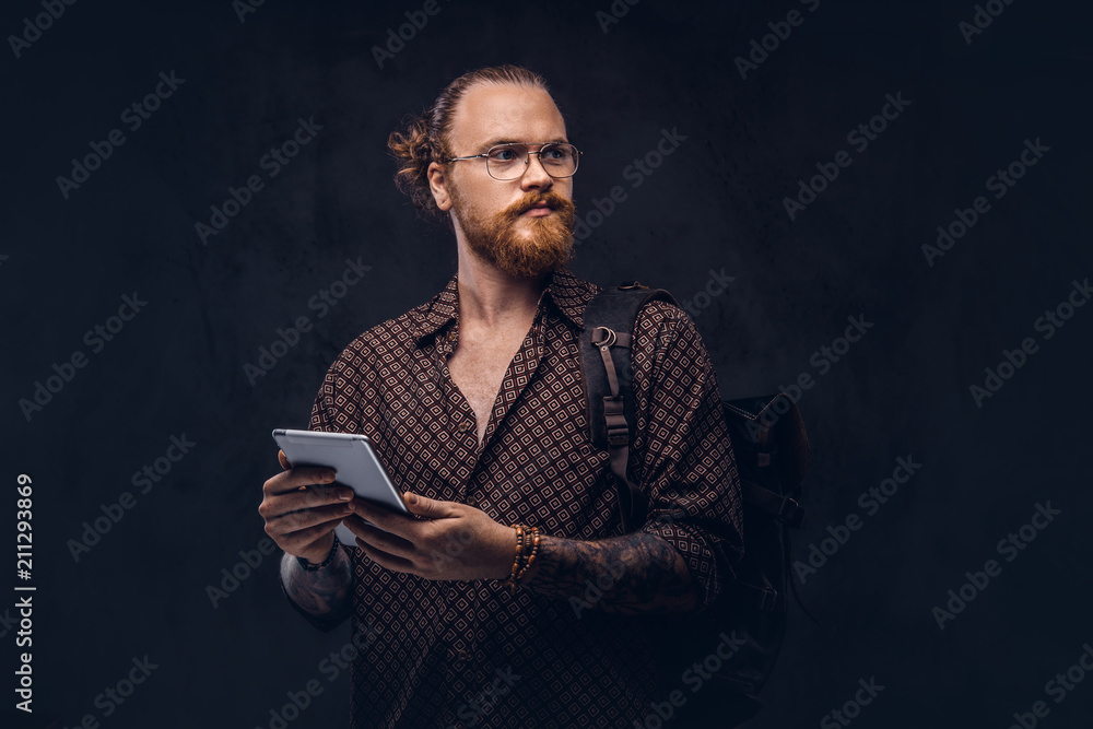 Portrait of a redhead hipster student in glasses dressed in a brown shirt, holds a backpack and digital tablet, posing at a studio. Isolated on a dark background. Portrait of a redhead hipster student in glasses dressed in a brown shirt, holds a backpack and digital tablet, posing at a studio. Isolated on a dark background.