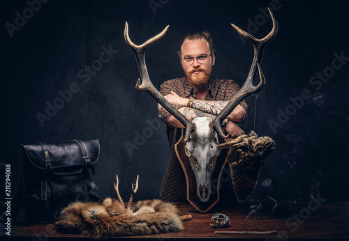 Redhead taxidermist hipster male in sunglasses dressed in a brown shirt, standing near a table with handmade trophy, owl scarecrow, and the fox skin. Isolated on the dark background. photo