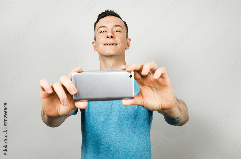 Young funny caucasian guy in blue t-shirt shoots on the smartphone's camera isolated on light gray background. Young funny caucasian guy in blue t-shirt shoots on the smartphone's camera isolated on light gray background.
