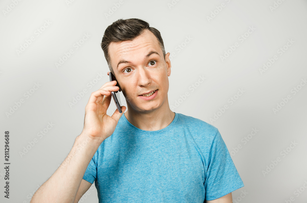 Young funny caucasian guy in blue t-shirt talking on the phone, isolated on light gray background. Young funny caucasian guy in blue t-shirt talking on the phone, isolated on light gray background.