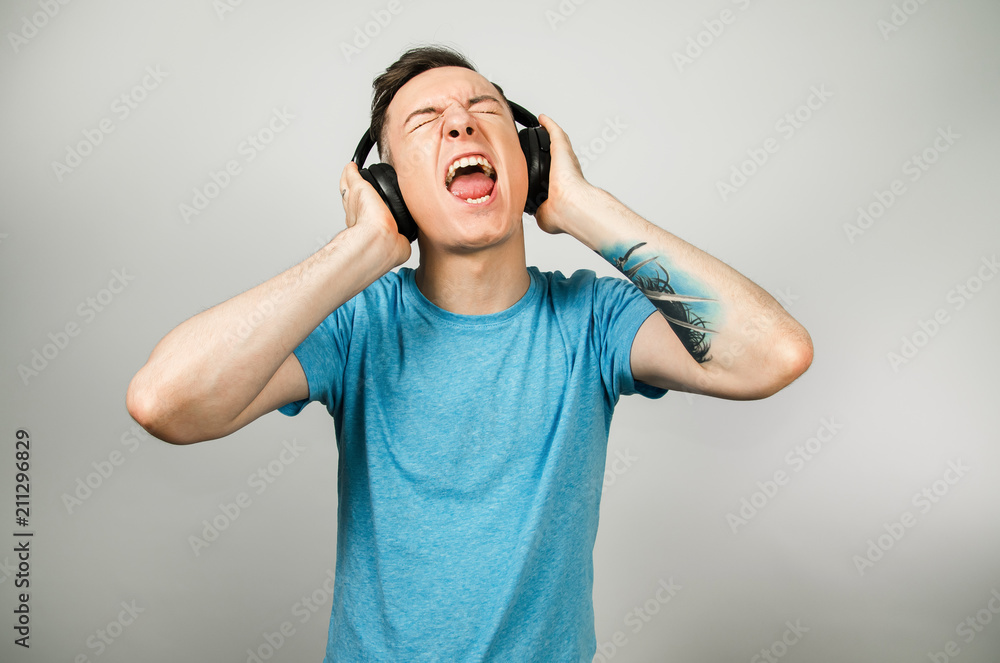 Young funny caucasian guy in blue t-shirt isolated listens to rock music on headphones on light gray background. Young funny caucasian guy in blue t-shirt isolated listens to rock music on headphones on light gray background.