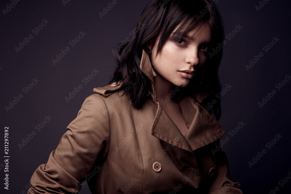 Dark portrait of beautiful sensual woman with nude makeup, dark hair, wearing beige trench coat. Studio shot Dark portrait of beautiful sensual woman with nude makeup, dark hair, wearing beige trench coat. Studio shot