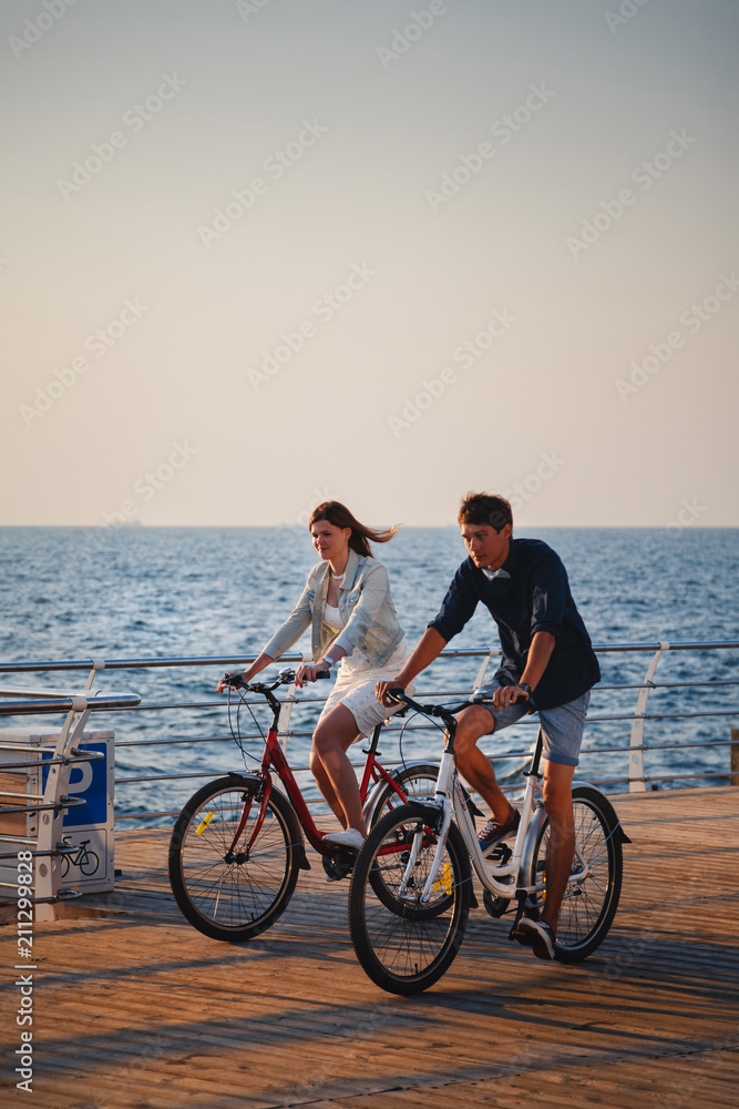 Couple of young hipsters cycling together at the beach at sunrise sky at wooden deck summer time