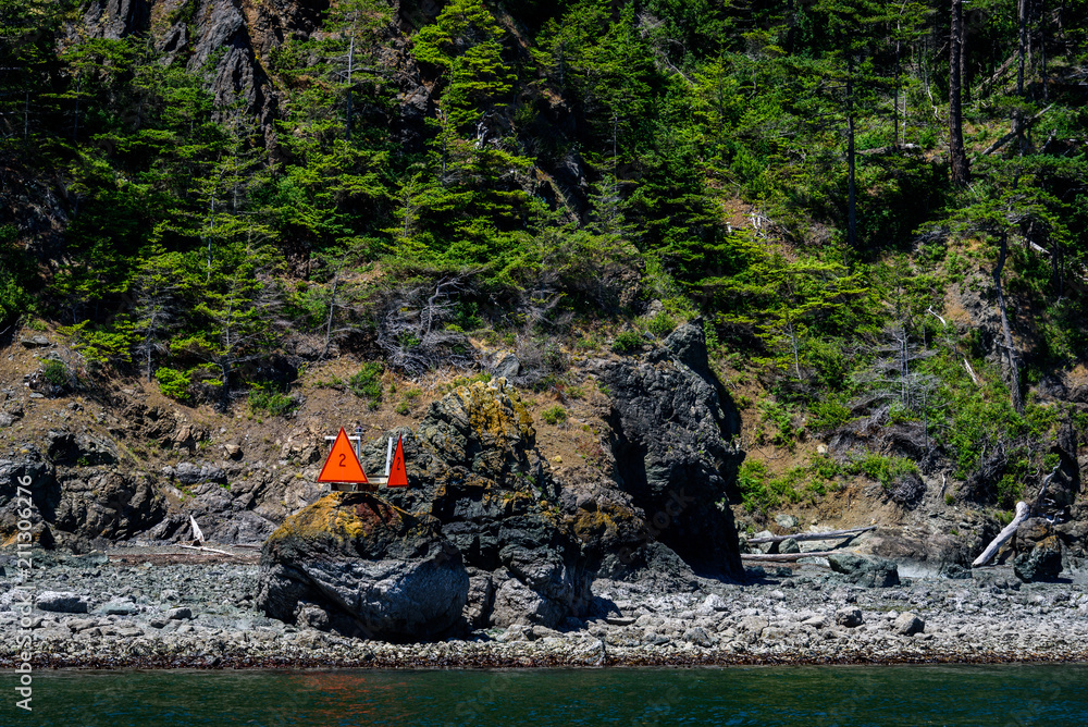 Rocky beach and tree covered hillside of a San Juan Island, orange hazard channel marker on a shoreline boulder, green water in the foreground
Rocky beach and tree covered hillside of a San Juan Island, orange hazard channel marker on a shoreline boulder, green water in the foreground