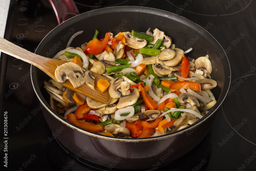 Stirring vegetables with mushrooms in a frying pan on a dark background. Cooking food Stirring vegetables with mushrooms in a frying pan on a dark background. Cooking food