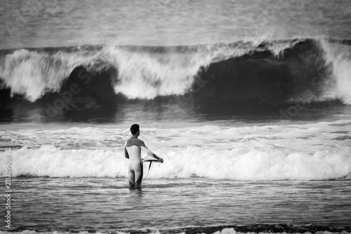 black and white sport surf picture with lonely surfer look a t a big waves in the background. wait the right time to enter in the dangerous water and have fun