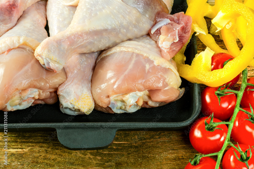 Raw organic chicken drumsticks with fresh vegetables lies in the black frying pan, on a wooden background before preparation. Cooking concept.