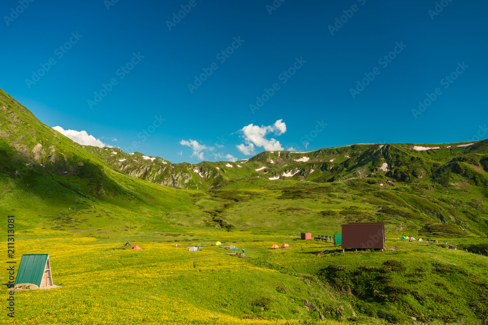 Mountain shelter on a green slope of the high big mountain. Alpine meadows in the Caucasus Mountains. Beautiful blue sky and clouds. Impressions from mountain tourism