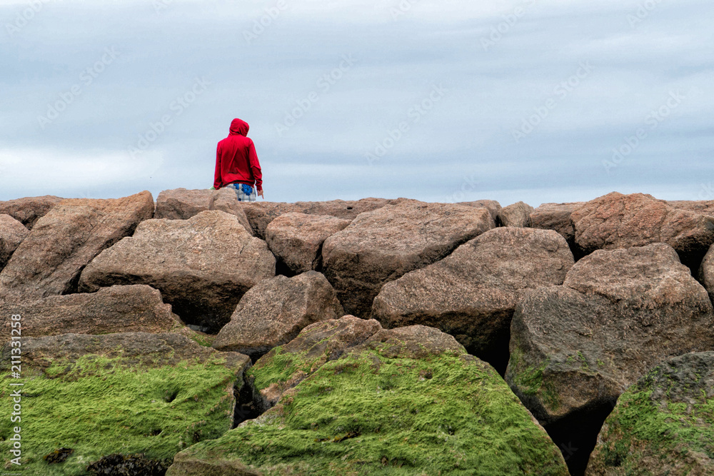 Man in a red hoodie standing beside rocks covered in seaweed looking out over a dismal sky.