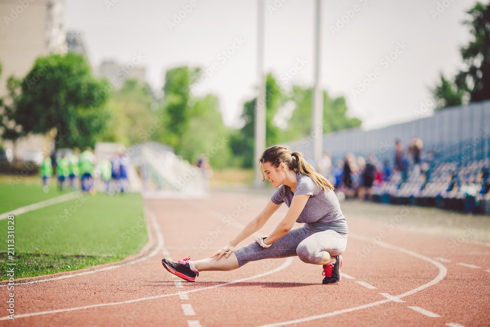 Beautiful young caucasian woman with long hair in tail and big breasts doing exercises, warming up and warming up muscles before training in running stadium, red rubber track in summer on a sunny day