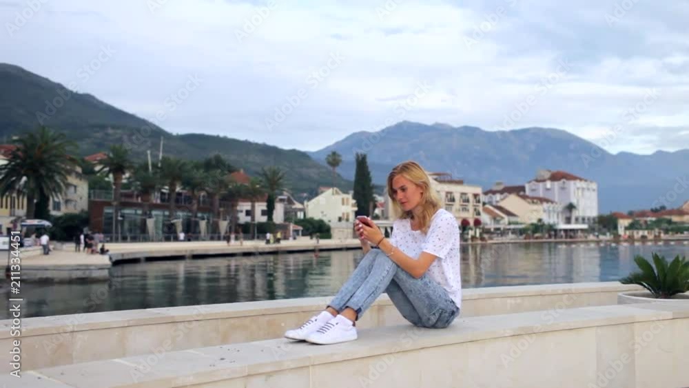 beautiful young woman with phone sits on the embankment of a luxury town beautiful young woman with phone sits on the embankment of a luxury town