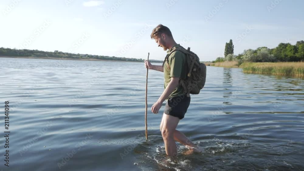 Young male with backpack and wooden hiking staff crossing a big river in summer sunny day Young male with backpack and wooden hiking staff crossing a big river in summer sunny day