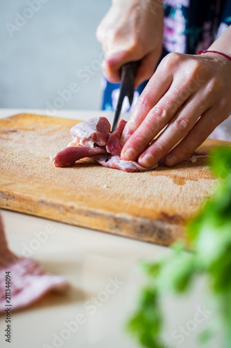 Woman hands cuts raw red meat into pieces with sharp knife on wooden board.