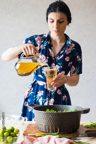 Woman hands adds white wine to chakapuli ingredients: lamb meat, green tkemali sour plum, onion sprouts, cilantro, tarragon leaves. Georgian traditional stew food photo