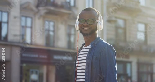 Portrait shot of the young African American smiled man in the headphones turning his head to the camera on the street of thetown on a sunny day. Outdoor. photo