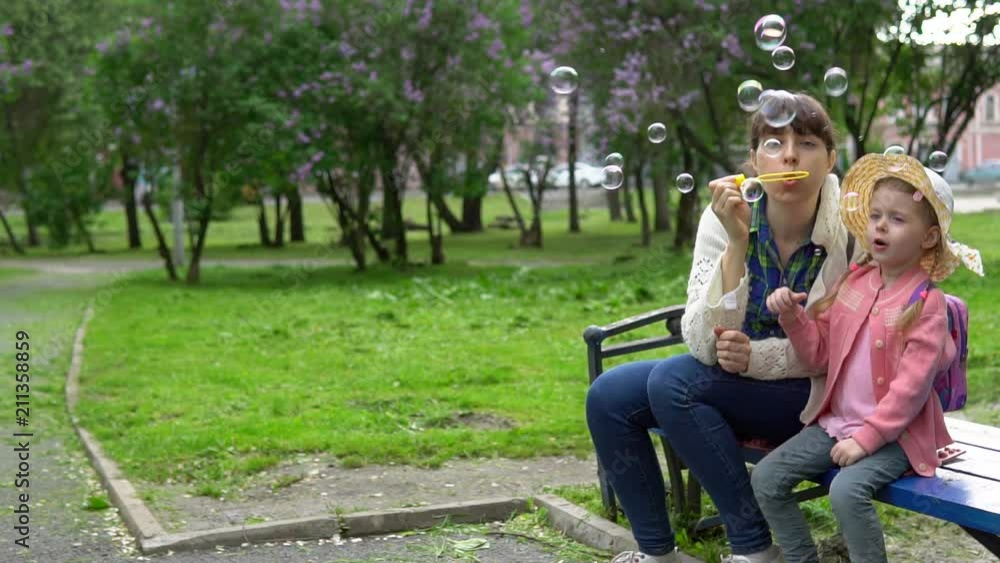 Young mother and cute daughter blowing soap bubbles in park of blooming lilacs on sunny spring day, slow motion. Young mother and cute daughter blowing soap bubbles in park of blooming lilacs on sunny spring day, slow motion.