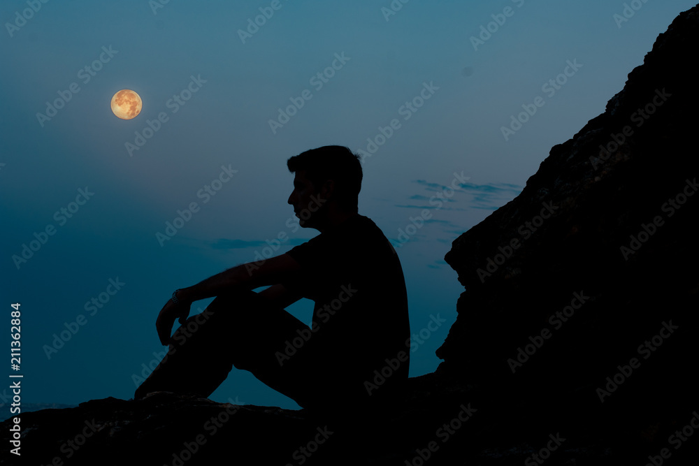 Foto de Silueta de hombre sentado en una roca de noche con luna do Stock