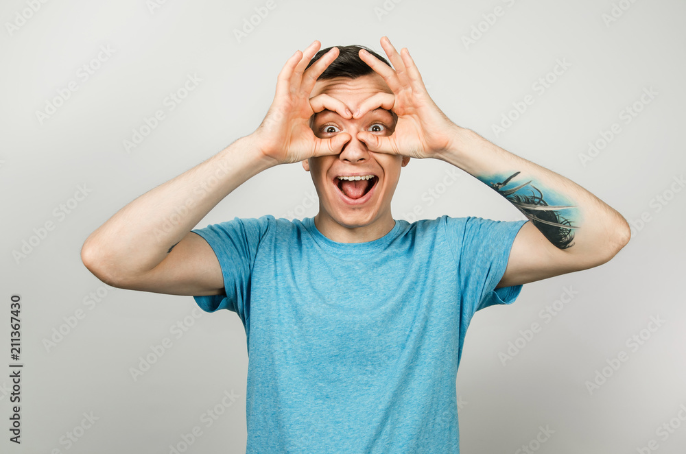 Young guy dressed in a blue t-shirt looks through binoculars from the palms of his hands, isolated on a light background. Young guy dressed in a blue t-shirt looks through binoculars from the palms of his hands, isolated on a light background.