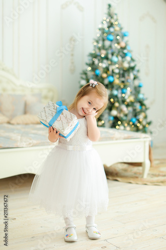 Little happy girl keeping present, wearing white dress and standing in bedroom with Christmas tree. Concept of winter holidays and gifts. photo