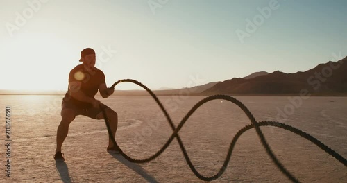 Slow motion: Athletic man doing crossfit battle rope workout outside at sunset in desert 