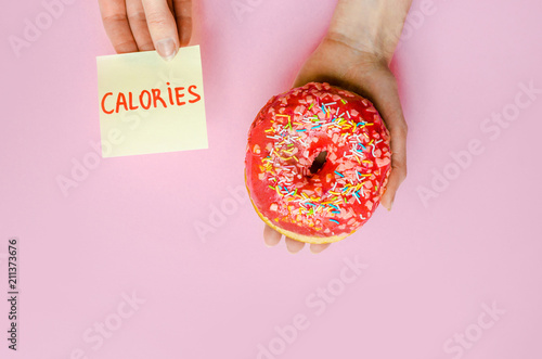 Woman hand holding pink donut with sprinkles extra calories fat fast food excess weight unhealthy on yellow background