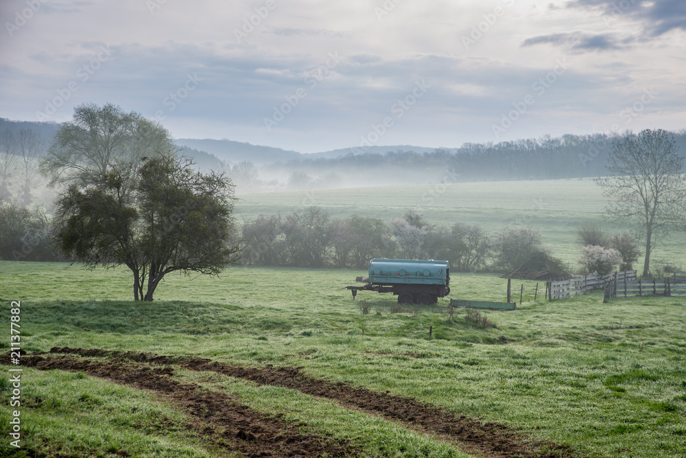 Fototapeta premium Trace d'un tracteur vers un pâturage avec son abreuvoir sur un paysage de brume à la campagne, avec au fond une forêt, près de Metz en Moselle