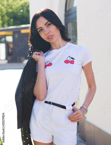 Summer sunny portrait of a beautiful brunette girl in a summer white outfit, shorts and t-shirt with cherries and a black jacket on her shoulder. Fashion. Street style.