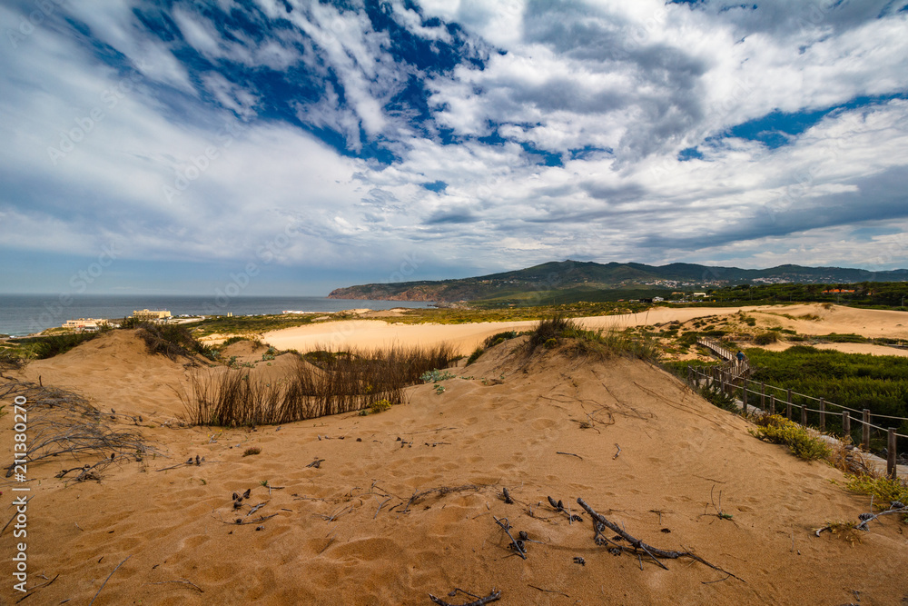 View of sand dunes on the Guincho beach near Atlantic coast. Landscape of sunny day, blue sky and a mountain in background. Cascais. Portugal. View of sand dunes on the Guincho beach near Atlantic coast. Landscape of sunny day, blue sky and a mountain in background. Cascais. Portugal.